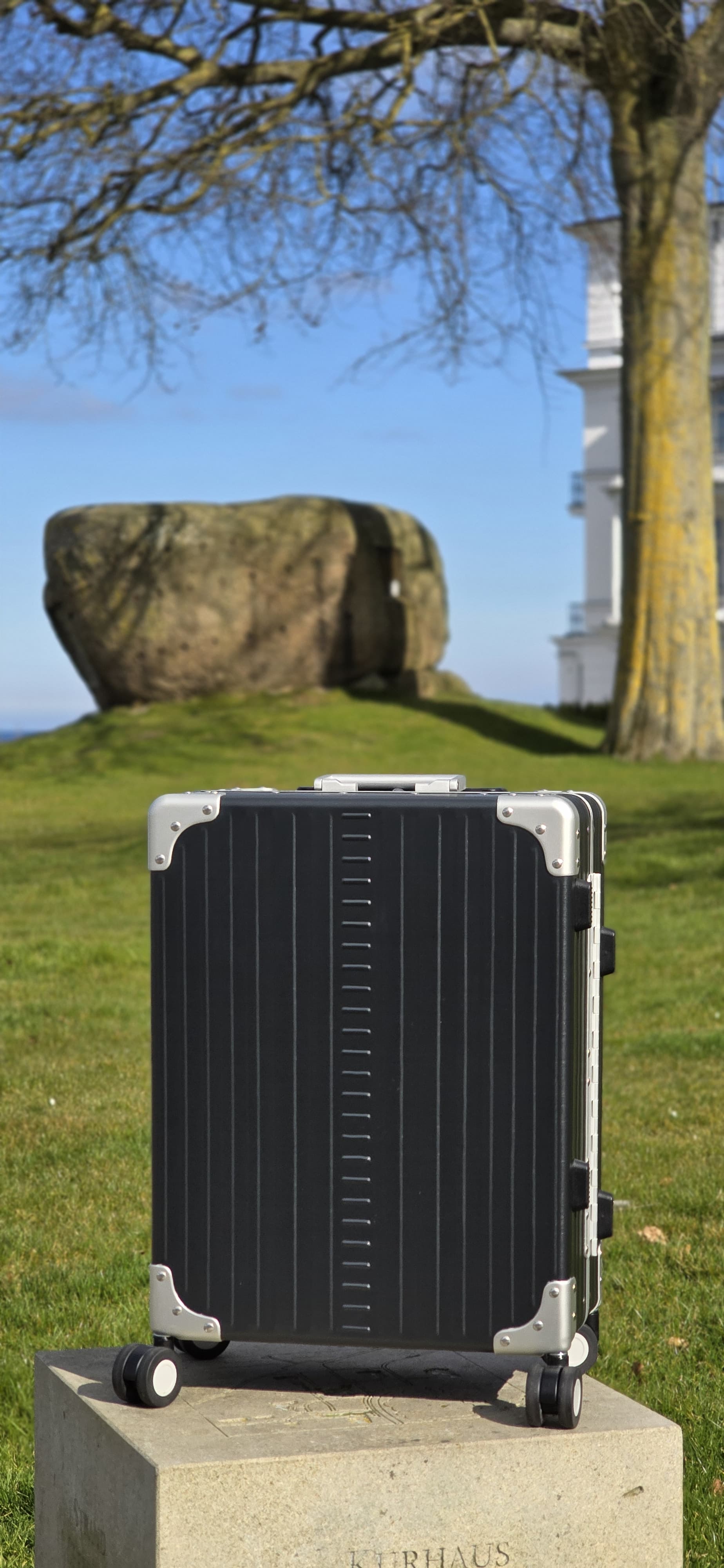 Valise cabine ALEON Fortis noire posée sur un socle en pierre devant un bâtiment blanc